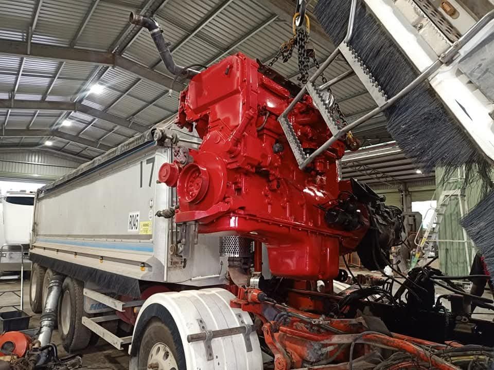 Red agricultural machinery engine block suspended from chains inside a workshop, with hoses and hydraulic components visible.