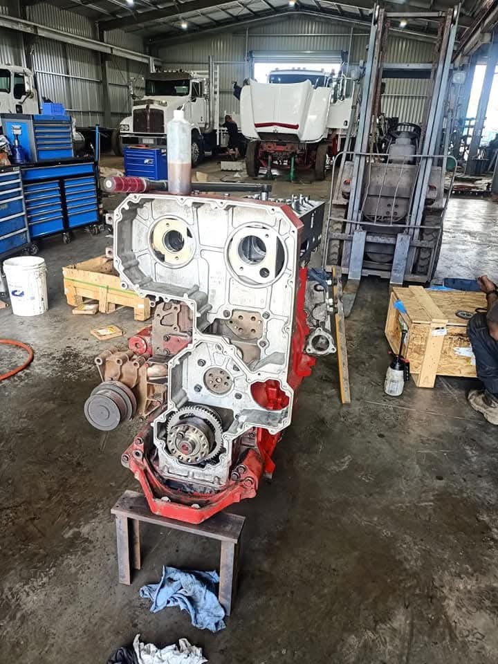 A stripped-down diesel truck engine sits in an agri repair workshop, surrounded by tool cabinets, workshop equipment and heavy vehicle parts during overhaul.
